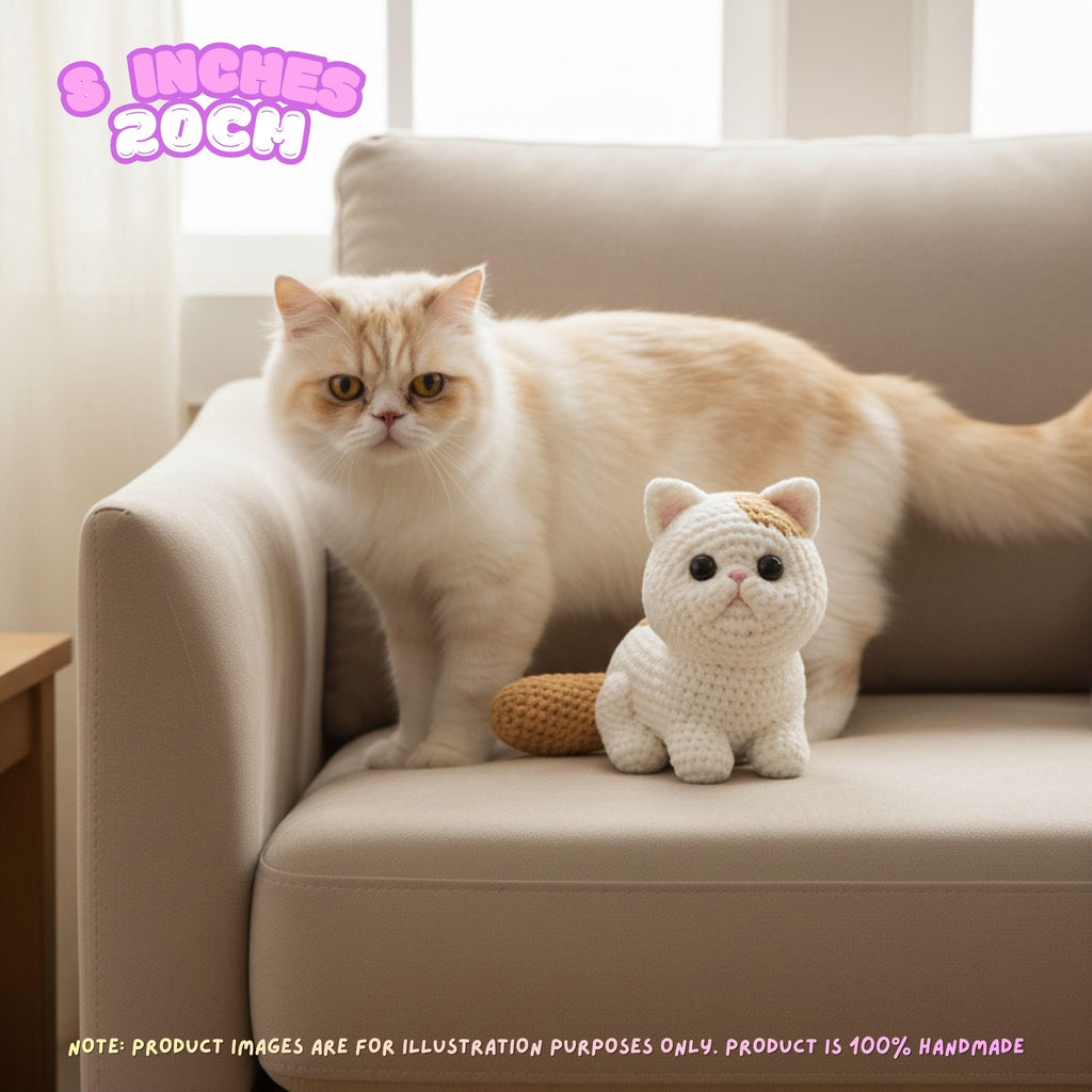 A cream-colored real cat stands on a beige sofa next to a small crocheted white cat toy. The scene is indoors with soft window light, and a pink text bubble in the top left says '8 INCHES 20CM'.