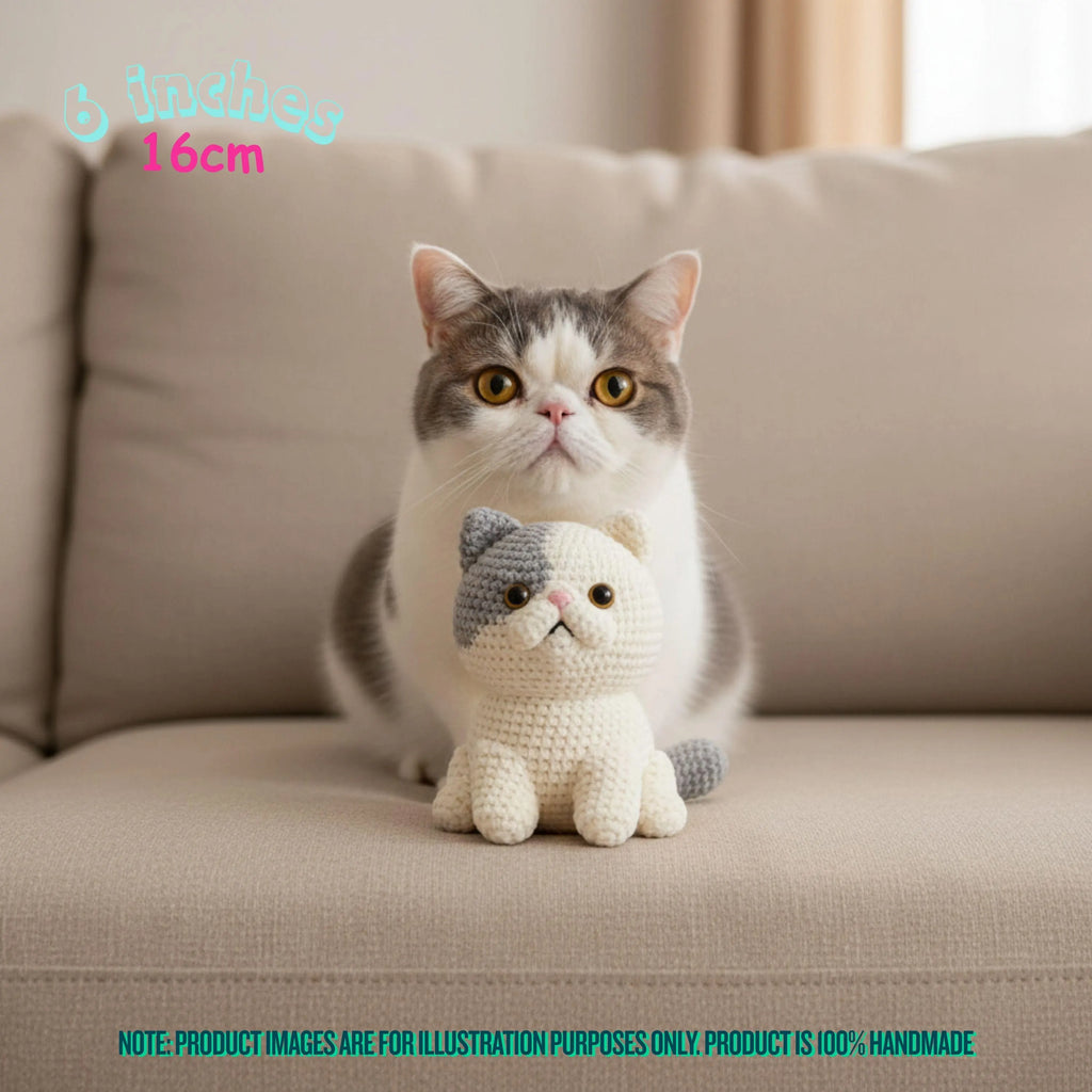 A real cat sits on a beige sofa next to a small crocheted cat toy. The cat is white and gray, and the toy is cream with gray ears and tail.
