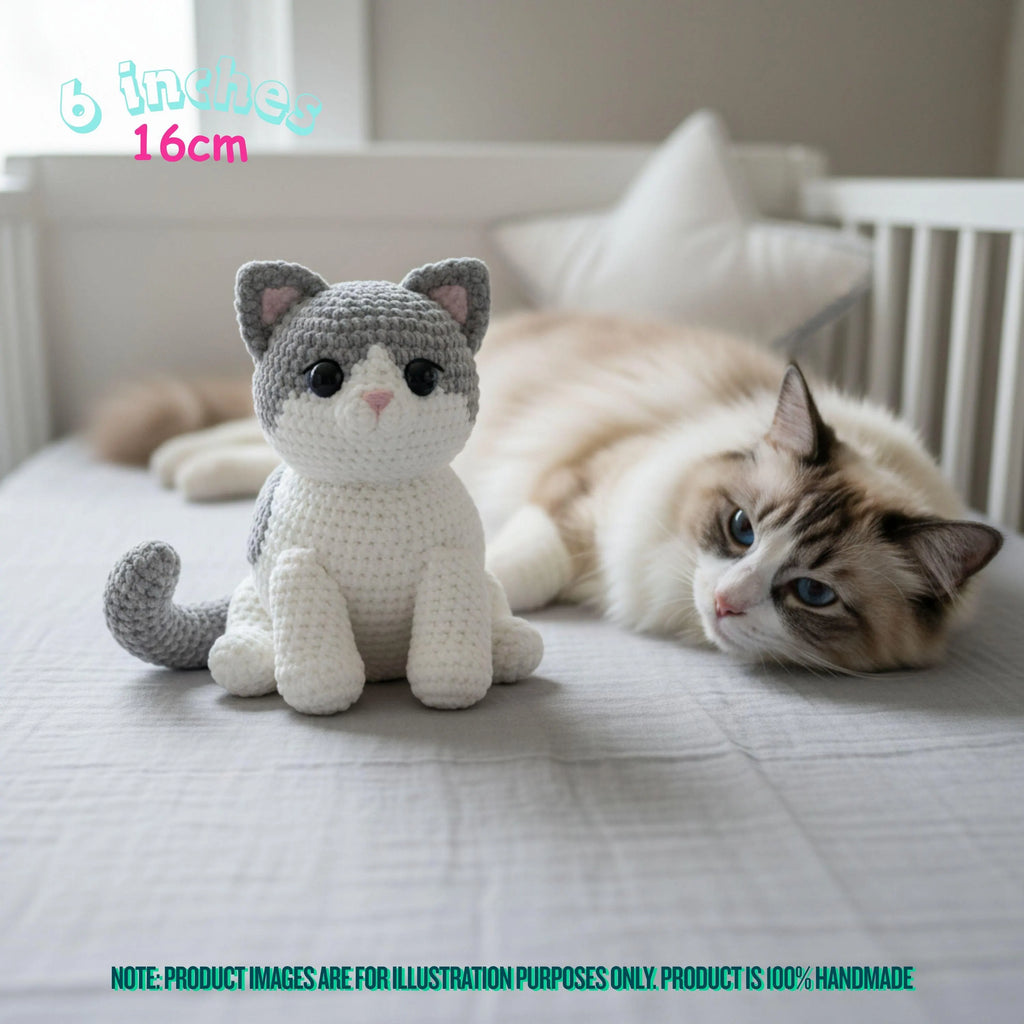 A crocheted cat plush toy sits on a bed next to a real fluffy cat in a nursery setting. Text overlays indicate the size is 6 inches / 16cm, with a note at the bottom stating product images are for illustration purposes only.