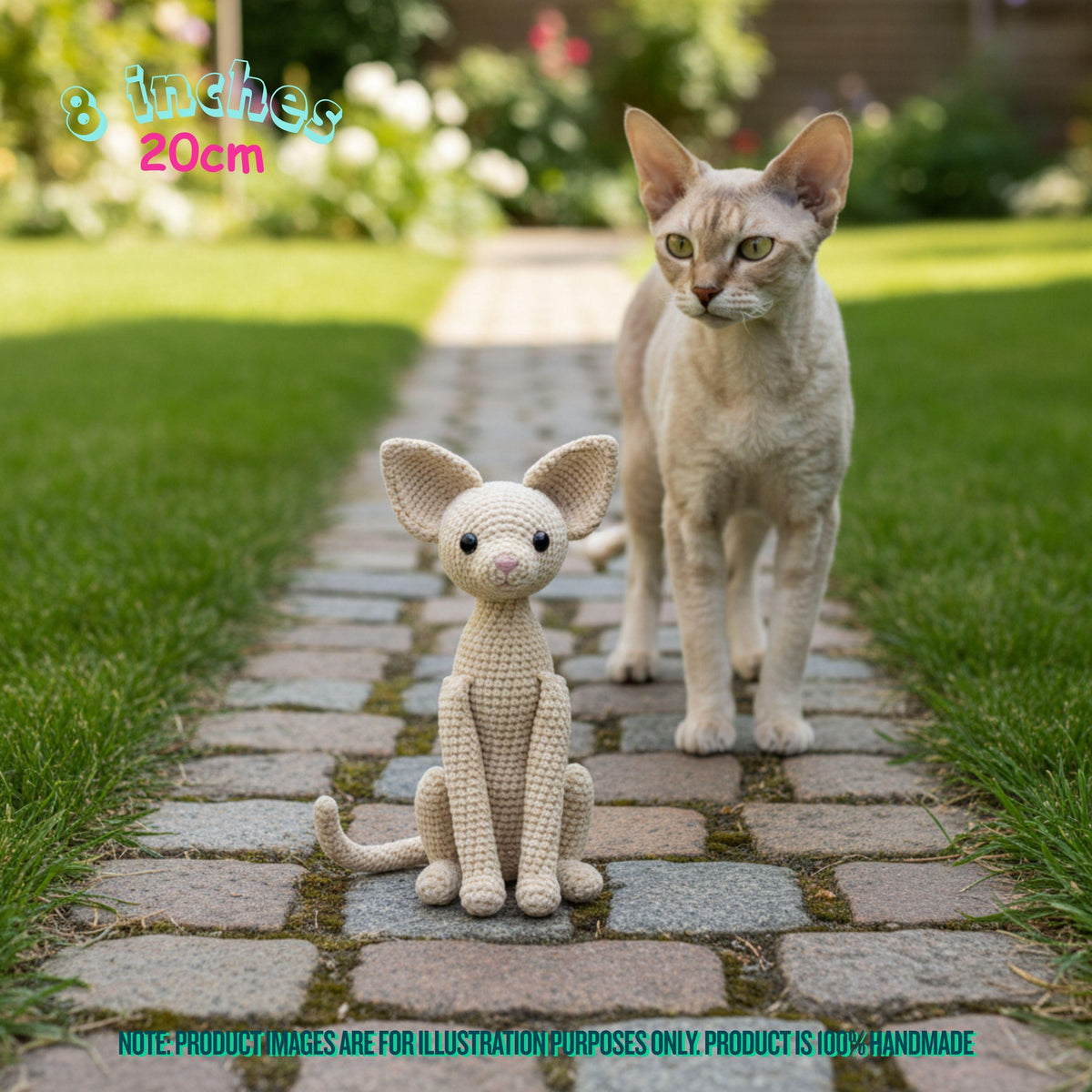 A crocheted cat toy sits on a stone garden path in front of a real cat standing on the same path. The scene is outdoors with grass and blurred garden plants in the background.