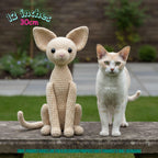 A crocheted cat-shaped plush toy sits on a stone ledge next to a real cat outdoors, with text in the top left reading '12 inches 30cm'. The toy is light cream/beige with black eyes and nose.