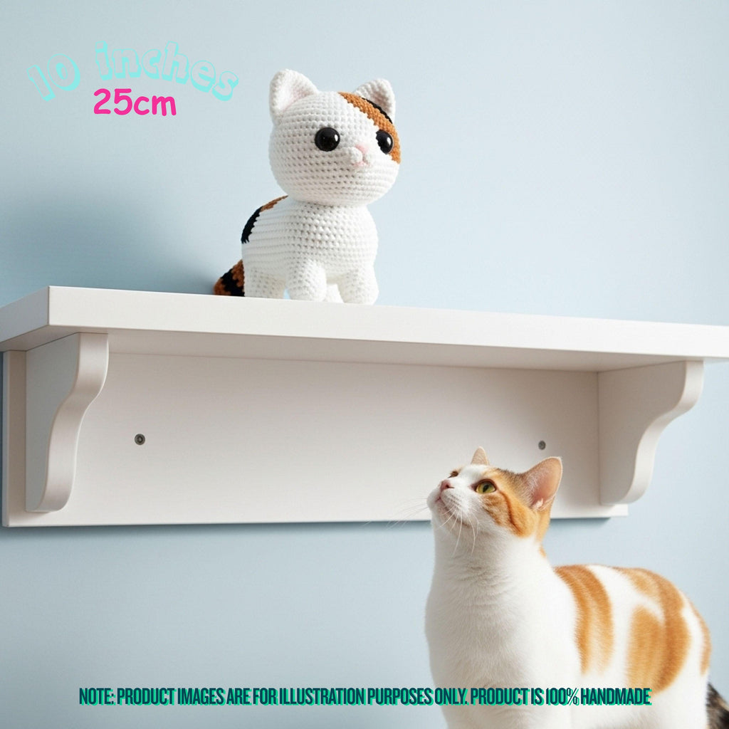 A white wall-mounted shelf with a crocheted calico cat toy displayed on top, with a real calico cat looking up at the shelf below. Text in the image indicates the shelf is 10 inches by 25 inches and notes the product is handmade.