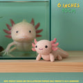 A crocheted axolotl toy in pale pink and coral pink is displayed on a wooden surface in front of a glass aquarium containing a real axolotl. Text in the top right reads '6 INCHES 15CM' and a note at the bottom says product images are for illustration purposes only.