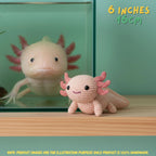 A crocheted axolotl toy in pale pink and coral pink is displayed on a wooden surface in front of a glass aquarium containing a real axolotl. Text in the top right reads '6 INCHES 15CM' and a note at the bottom says product images are for illustration purposes only.