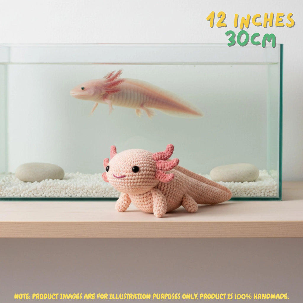 A crocheted axolotl toy in pale pink and peach sits on a wooden shelf in front of a glass aquarium with white gravel and a real axolotl swimming inside. Yellow text in the top right reads '12 INCHES 30CM', and a note at the bottom says the product images are for illustration purposes only because the product is 100% handmade.