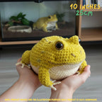 A yellow and cream crocheted frog toy being held in two hands, with a real indoor environment and a glass terrarium visible in the background. Text overlays indicate the size as 10 inches (25 cm) and note that the product image is for illustration purposes since the product is 100% handmade.