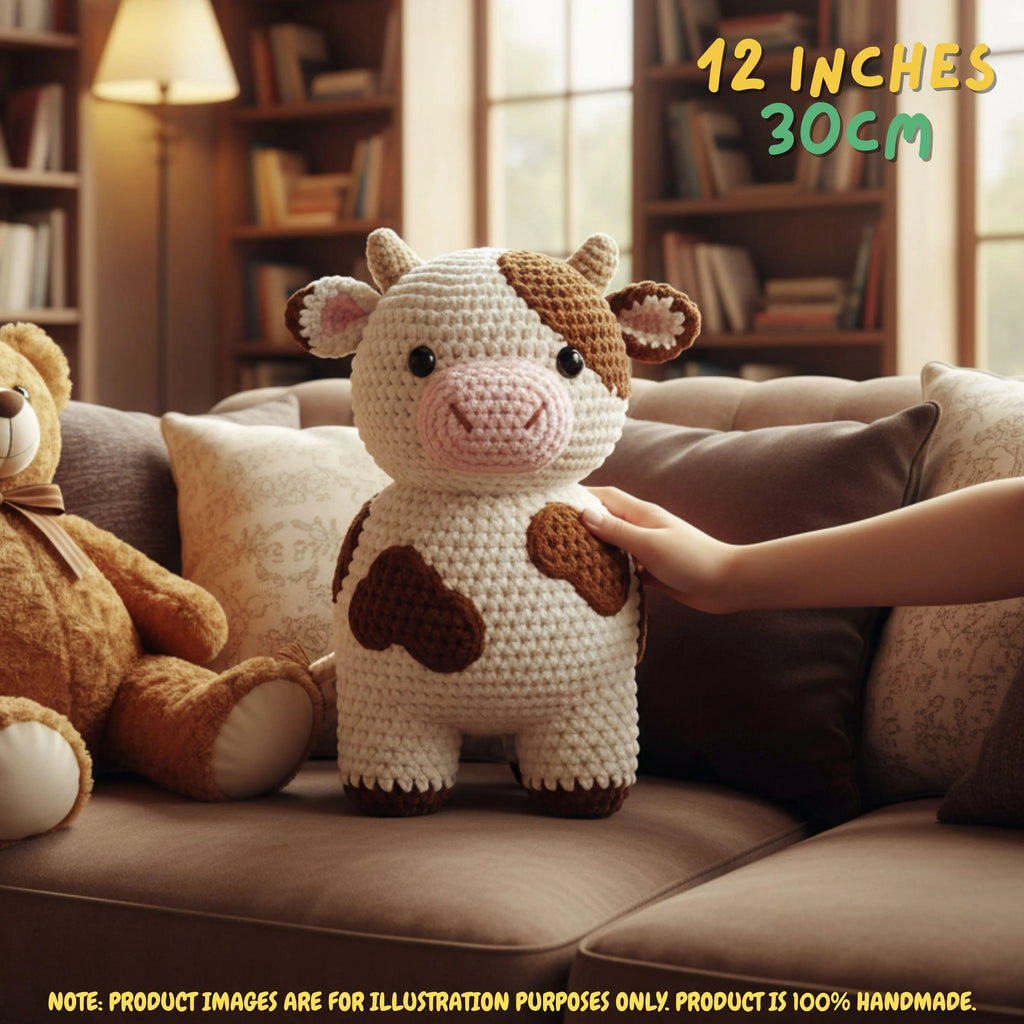 A crocheted cow plush toy sits on a brown sofa in a cozy living room, with a human hand touching its side. A teddy bear is partly visible on the left, and text in the top right reads '12 INCHES 30CM'.