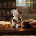 A crocheted cow plush toy sits on a wooden table beside an open book and a hand holding a cup of coffee. The background shows a softly blurred bookshelf, and text in the top right reads '6 INCHES 16CM'.