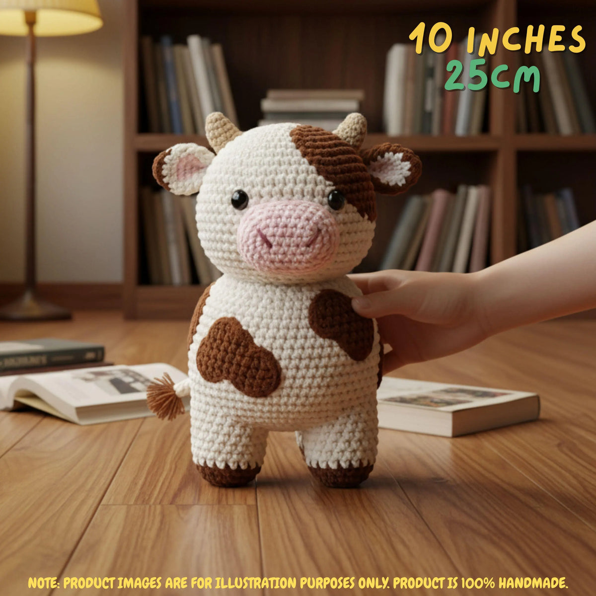 Crocheted cow plush toy standing on a wooden floor in front of a bookshelf, with a human hand touching its side. Text overlays read '10 INCHES 25cm' and 'NOTE.
