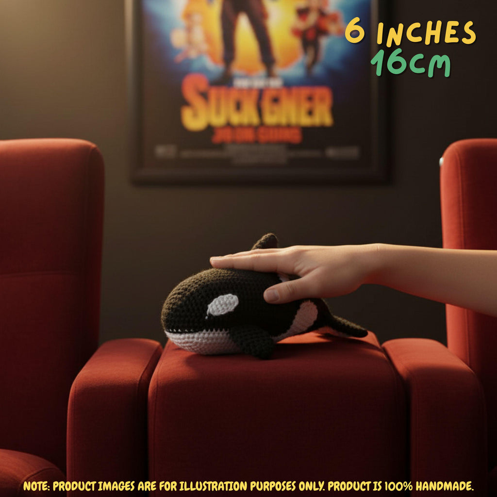 A crocheted orkala plush toy resting on a red theater-style chair, with a human hand gently touching its head. The background shows a movie poster for 'Sucker Punch' and text overlays indicating the size is 6 inches (16 cm).