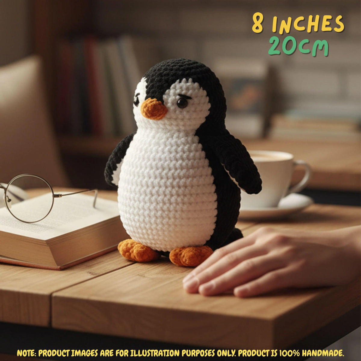 A crocheted penguin toy sits on a wooden table in a cozy indoor setting, with a hand resting near it, a book and glasses nearby, and a cup in the background. Text in the top right reads '8 INCHES 20cm', and a note at the bottom says product images are for illustration purposes only.