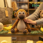 A crocheted monkey plush toy sits on a glass table in a warm, cozy shop setting. A person's hand gently touches the monkey's back, and other handmade items like a crocheted tote bag, yarn, and a teddy bear are visible in the background.