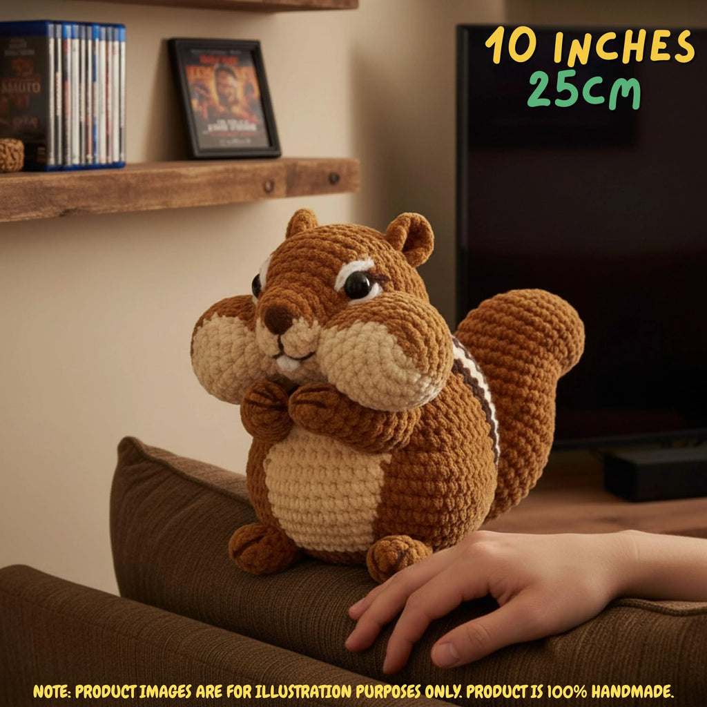 A crocheted squirrel plush toy sitting on a brown couch in a living room setting, with a human hand resting near it. The image includes text overlays indicating the size as 10 inches / 25 cm and a note that the product image is for illustration purposes only.