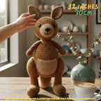 A crocheted kangaroo plush toy sits on a wooden table in a cozy indoor setting, with a human hand touching its head. The plush is made of brown and beige yarn and has embroidered facial features.