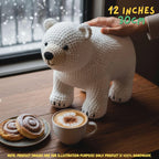 A white crocheted polar bear figurine standing on a wooden table by a window, with a hand resting on its back. A plate with cinnamon rolls and a cup of latte are in the foreground, and text in the top right says '12 INCHES 30CM'.