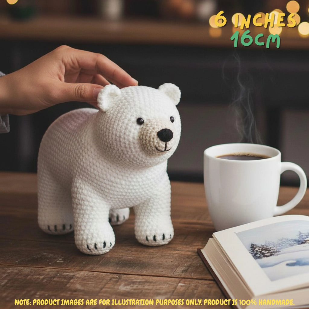 A crocheted white polar bear toy sits on a wooden table next to a white mug of coffee and an open book. A human hand is gently touching the bear's back.
