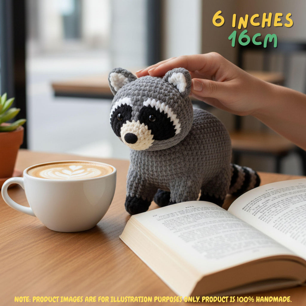 A crocheted raccoon plush toy sits on a wooden table next to an open book and a cup of latte. A human hand is gently touching the raccoon's head.