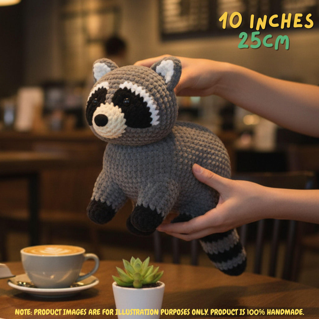 A crocheted raccoon plush toy being held by two hands in a cozy indoor setting, with a coffee cup and small succulent on a table in the foreground. Text overlays indicate the size as 10 inches (25cm) and note that the product is 100% handmade.