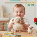 A baby sitting at a table holds a crocheted sheep toy while another smaller crocheted sheep sits on the table. The scene is indoors with a blurred chair in the background, and text in the top right reads '8 INCHES 20CM'.