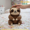 Handmade brown crochet sloth amigurumi doll sitting on a floral bed with a hand gently resting on its head, featuring "10 INCHES / 25CM" text overlay.