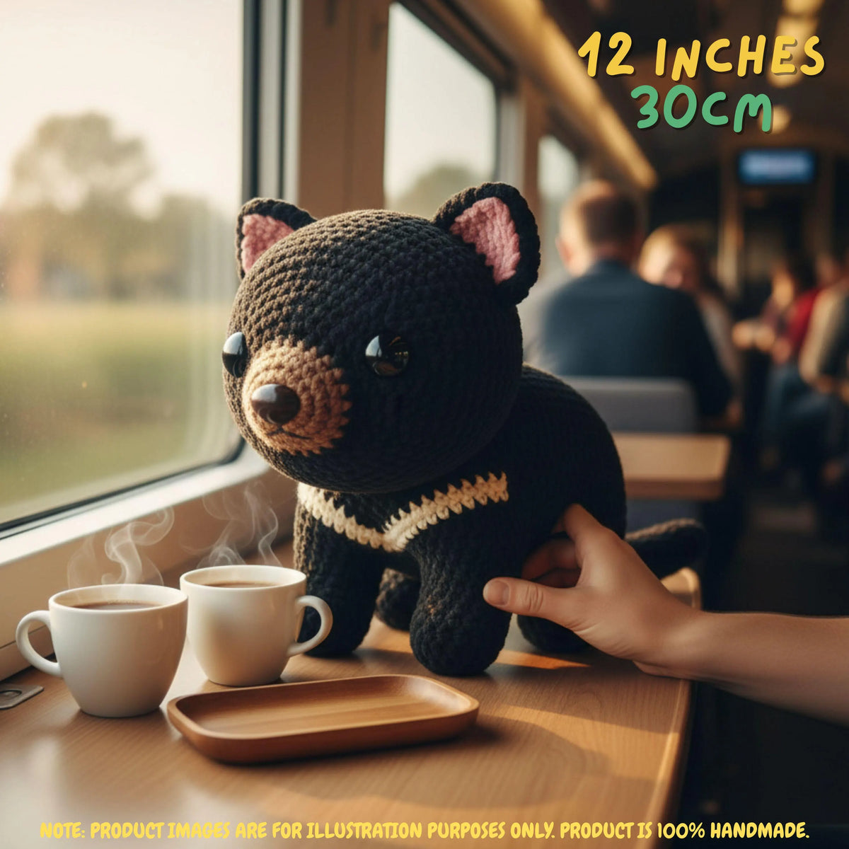 A crocheted black bear plush toy with pink inner ears, a brown snout, and a cream stripe, sitting on a train table next to two white mugs. A human hand is touching the plush, and text in the corner says 12 inches / 30 cm.