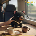 A crocheted black bear plush toy with pink inner ears and a cream-and-black striped neck band sits on a wooden table inside a train or vehicle. A human hand is holding the toy, and coffee mugs are in the foreground.