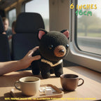 A crocheted black bear plush toy with pink inner ears and a cream-and-black striped neck band sits on a wooden table inside a train or vehicle. A human hand is holding the toy, and coffee mugs are in the foreground.