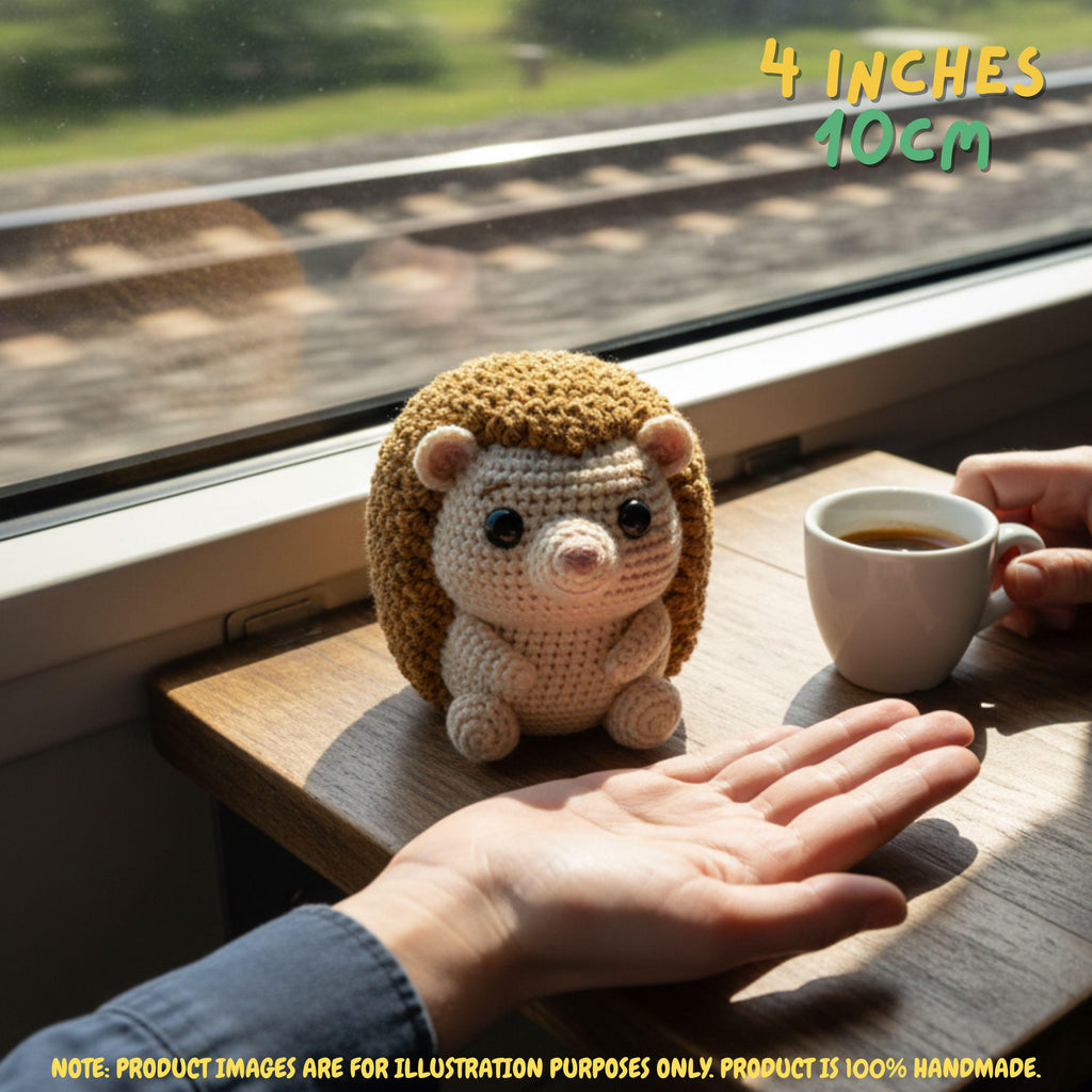 A crocheted hedgehog toy sits on a wooden windowsill next to a cup of coffee, with a person's hand reaching toward it. The background shows a blurred train window and tracks.