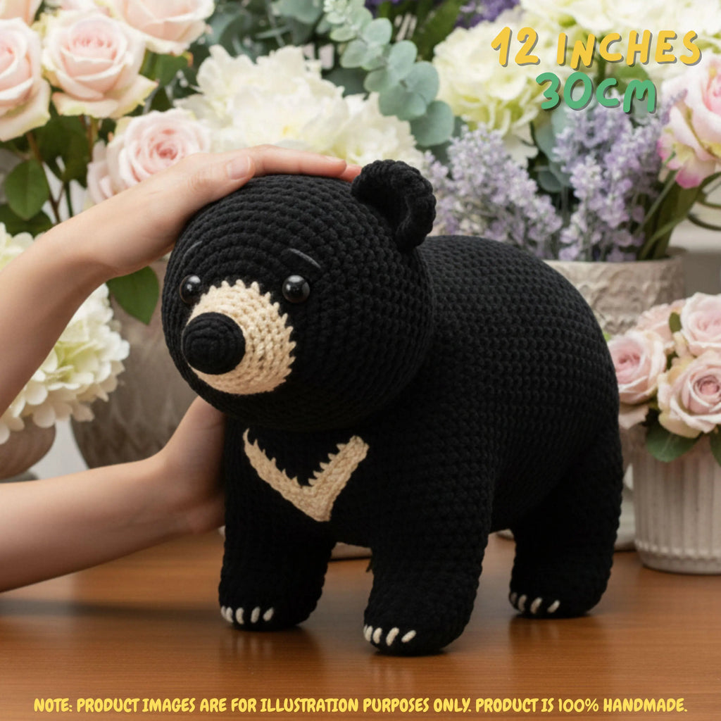 A crocheted black bear plush toy with beige facial and chest details, being held by a person's hand on a wooden table with a blurred floral background. Yellow text in the top right reads '12 INCHES 30CM'.