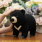 A crocheted black bear plush toy with beige facial and chest details, being held by a person's hand on a wooden table with a blurred floral background. Yellow text in the top right reads '12 INCHES 30CM'.