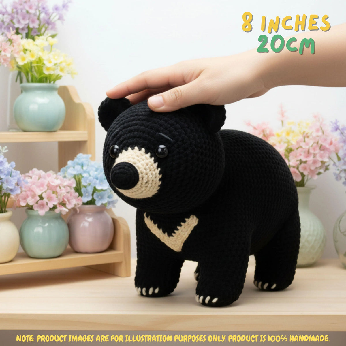 A crocheted black bear plush toy with cream-colored accents, shown with a human hand resting on its head. The background is a soft, blurred indoor setting with pastel flowers and shelves.