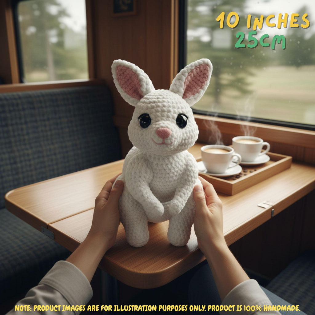 A white crocheted bunny plush toy with pink inner ears is held by two hands on a wooden table inside a train or bus cabin. A tray with two steaming cups sits nearby, and text in the top right reads '10 INCHES 25CM'.