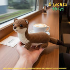 Handmade Stoat crochet amigurumi plush doll in brown and cream with a dark-tipped tail held in a hand on a wooden table, next to a coffee cup by a window, with text indicating 6 inches 16 cm size.