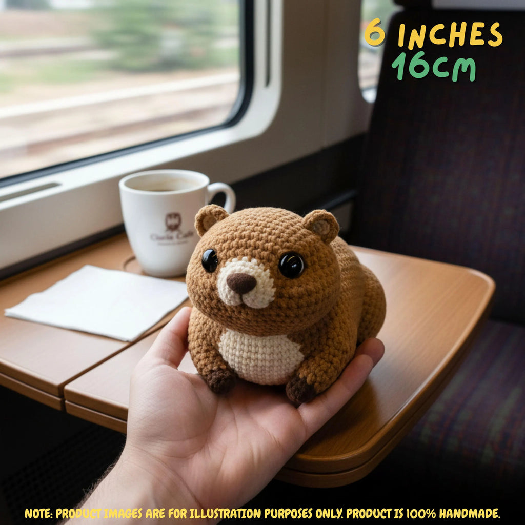 A small crocheted bear plush toy being held in a person's hand inside a train compartment. The bear is brown with a cream-colored chest and snout, black eyes, and small dark paws.