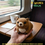 A small crocheted bear plush toy being held in a person's hand inside a train compartment. The bear is brown with a cream-colored chest and snout, black eyes, and small dark paws.