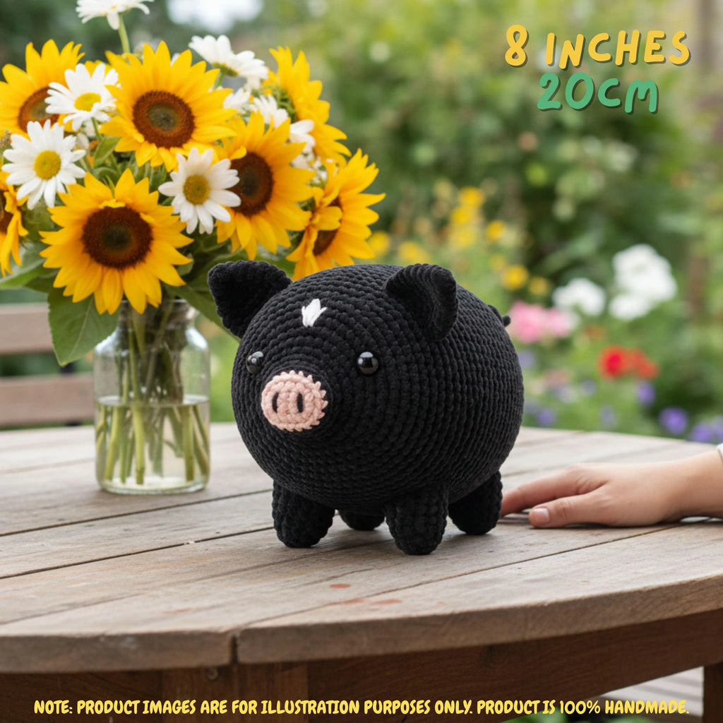 A black crocheted pig toy sits on a wooden table outdoors, with a hand resting near it and a vase of sunflowers in the background. Text in the top right reads '8 INCHES 20CM'.
