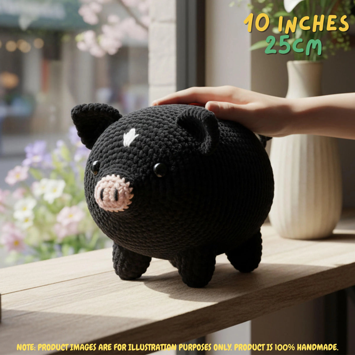 A black crocheted pig toy with a pink snout and white forehead detail, shown on a wooden surface with a human hand resting on its back. The background is a softly blurred indoor setting with flowers and a vase.