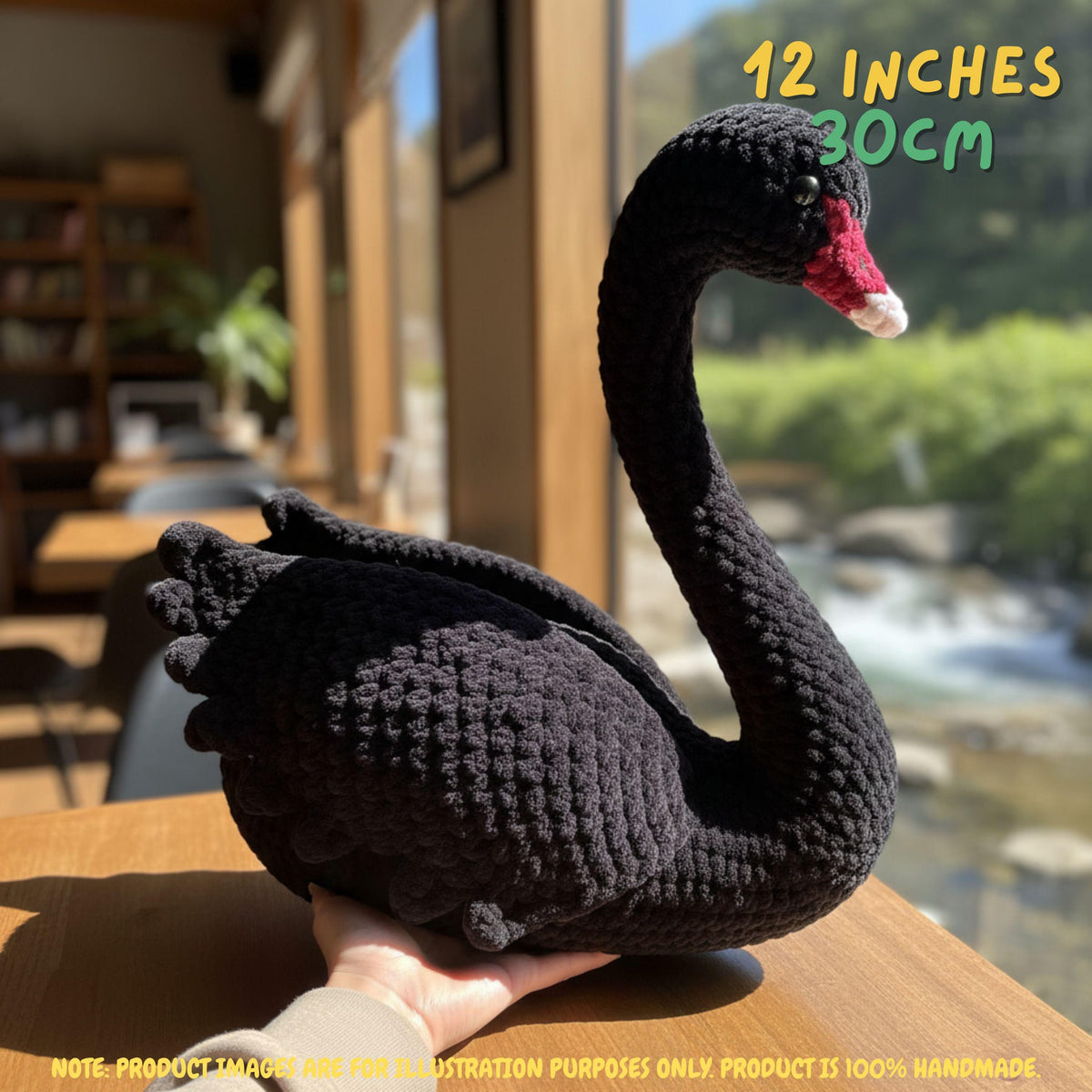 A crocheted black swan figurine with a red and white beak, held on a wooden table in a cafe setting with a blurred window view of a river and mountains in the background. Text overlays indicate its size as 12 inches (30cm) and note that the product is 100% handmade.