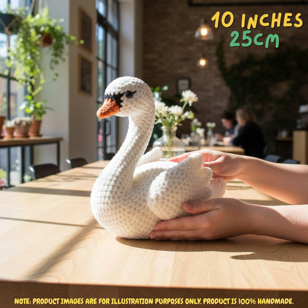 Handmade Mute Swan crochet amigurumi plush doll resting on a wooden table held gently by hands, with text indicating 10 inches 25 cm size in a bright cafe or room setting.