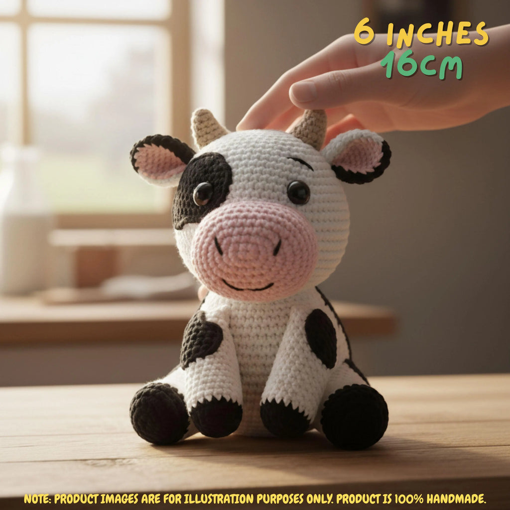 A crocheted cow plush toy sits on a wooden table with a human hand gently touching its head. The toy is white with black patches, a pink snout and inner ears, and small tan horns.