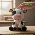 A crocheted cow plush toy sits on a wooden table with a human hand gently touching its head. The toy is white with black patches, a pink snout and inner ears, and small tan horns.