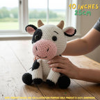 A crocheted cow plush toy sits on a wooden table in front of a window, with a person's hands holding it and text in the corner reading '10 INCHES 25CM'.