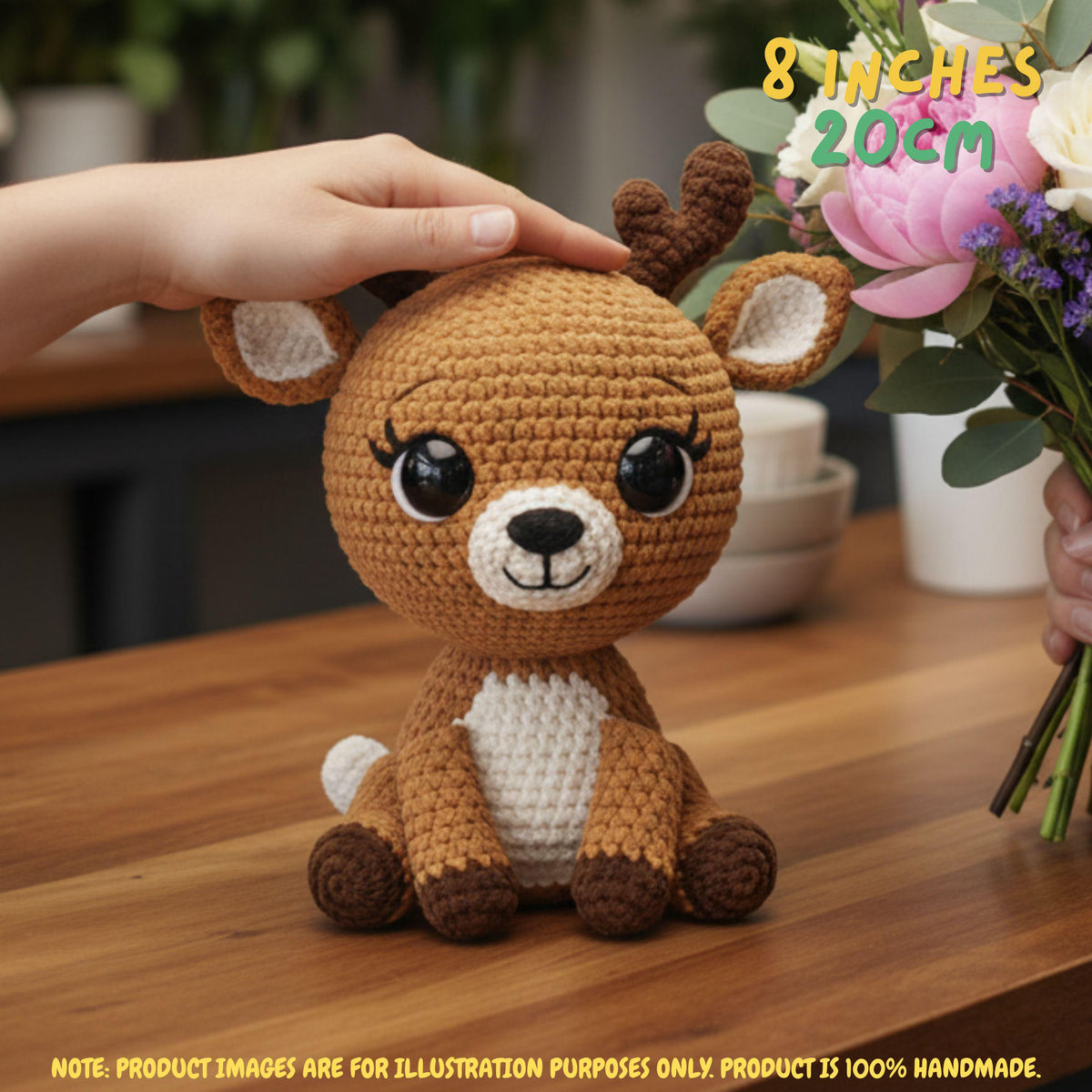 A crocheted stuffed deer toy sits on a wooden table, with a human hand resting on its head and flowers at the right edge. The image includes text overlays stating '8 INCHES 20CM' and a note about the product being 100% handmade.