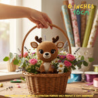 A crocheted stuffed deer toy sits in a woven basket filled with pink flowers, white daisies, and green leaves. A human hand holds the basket handle, and the scene is styled like a craft room with yarn spools and fabric rolls in the background.