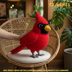 A red crocheted cardinal bird toy or plush is displayed on a round cushion inside a wicker chair, with a human hand supporting it. The setting is indoors with plants and warm lighting, and text in the top right says '12 INCHES 30cm'.