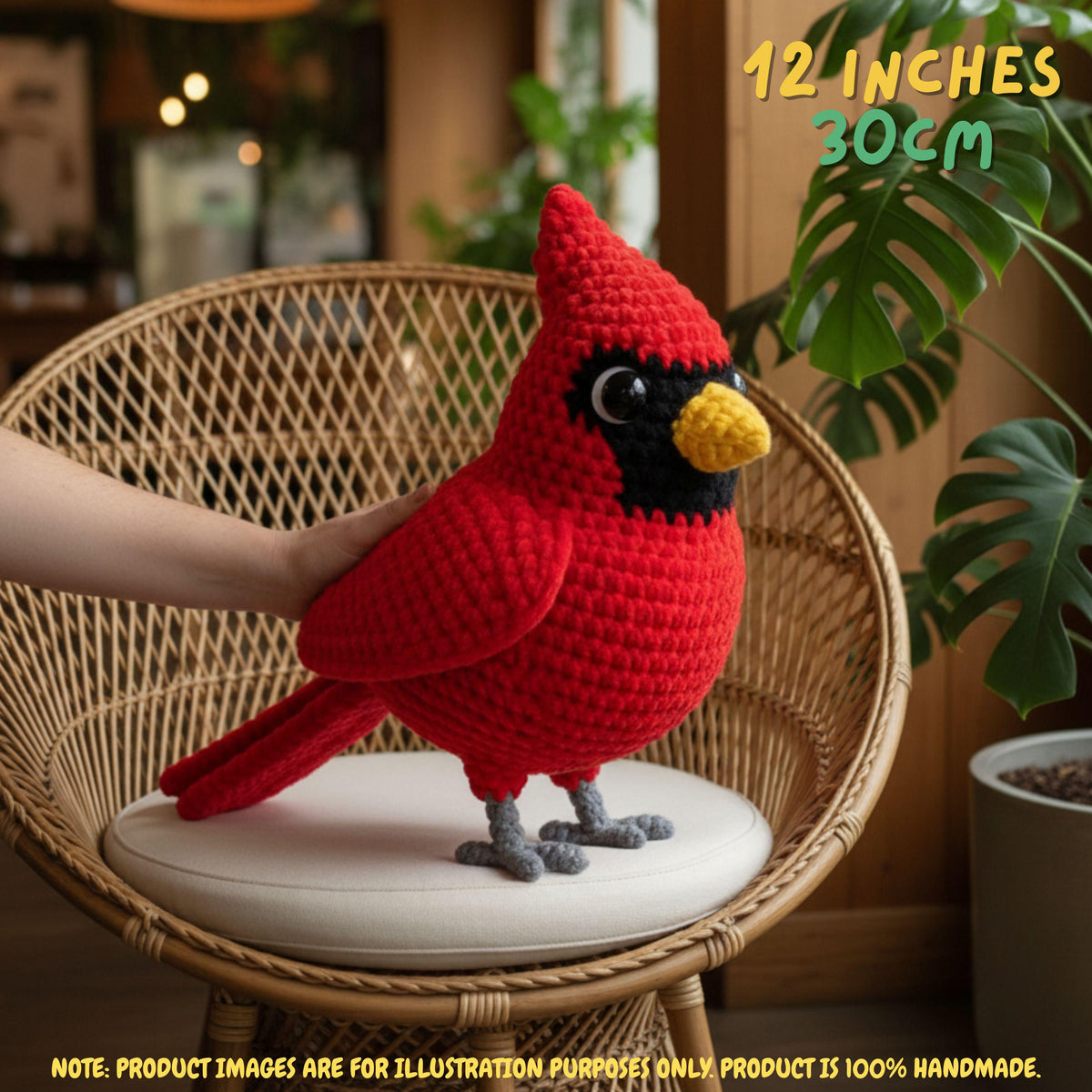 A red crocheted cardinal bird toy or plush is displayed on a round cushion inside a wicker chair, with a human hand supporting it. The setting is indoors with plants and warm lighting, and text in the top right says '12 INCHES 30cm'.