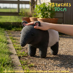 A crocheted animal-shaped plush toy with a dark gray head and legs and a white body, shown outdoors on a gravel path with a human hand resting on its back. Text in the top right reads '12 INCHES 30cm', and a note at the bottom says product images are for illustration purposes only.