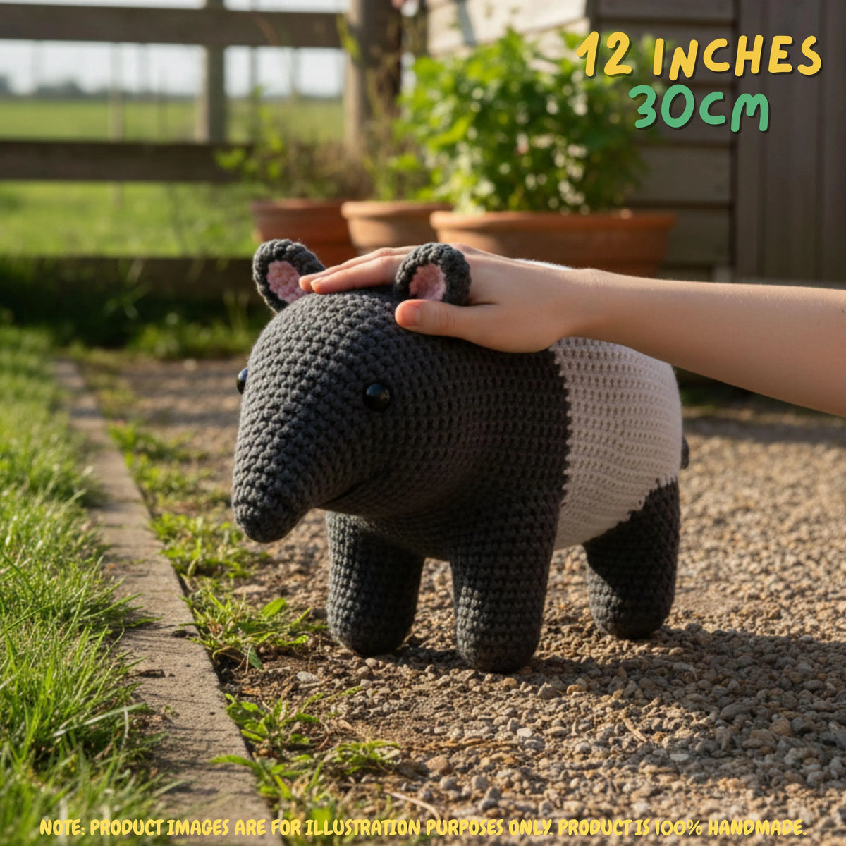 A crocheted animal-shaped plush toy with a dark gray head and legs and a white body, shown outdoors on a gravel path with a human hand resting on its back. Text in the top right reads '12 INCHES 30cm', and a note at the bottom says product images are for illustration purposes only.