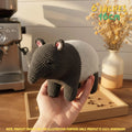 A crocheted stuffed animal resembling a small mammal with a dark gray head and legs and a white body, being held in a person's hand. The background shows a warm indoor setting with a coffee machine, dried flowers in a vase, and a wooden tray.
