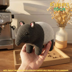 A crocheted stuffed animal resembling a small mammal with a dark gray head and legs and a white body, being held in a person's hand. The background shows a warm indoor setting with a coffee machine, dried flowers in a vase, and a wooden tray.